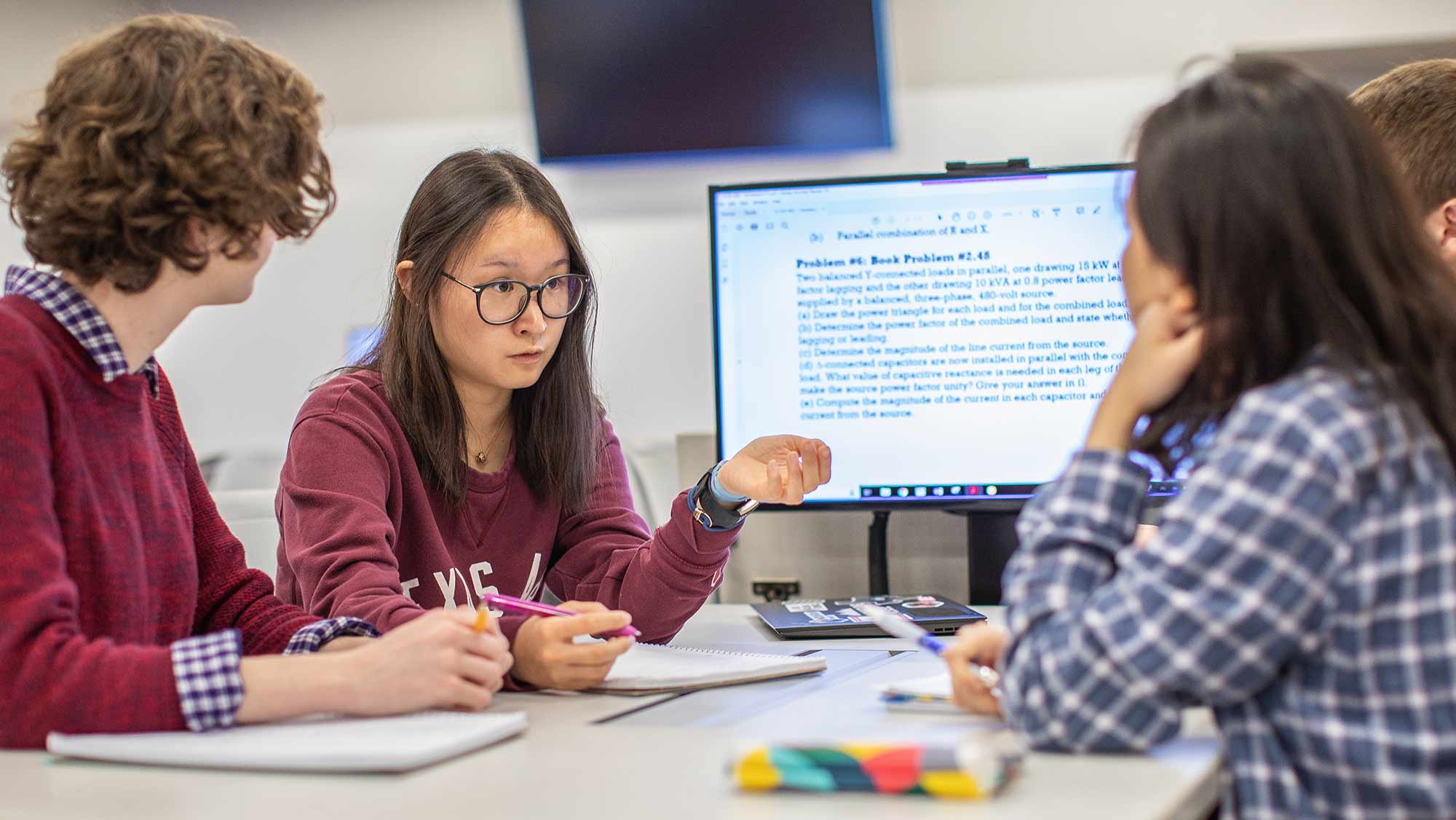 Four students talking and sitting at a classroom desk with a large screen at one end.