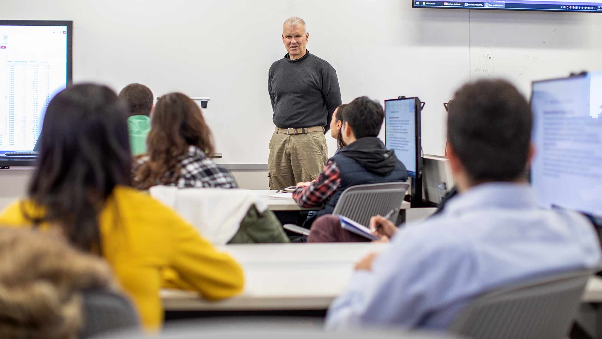 Professor talking at front of a classroom full of students sitting in chairs.