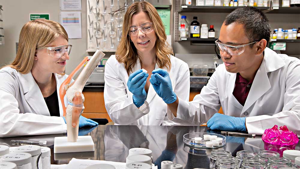 两个女人和一个男人穿着白大褂，坐在实验室的一张桌子旁