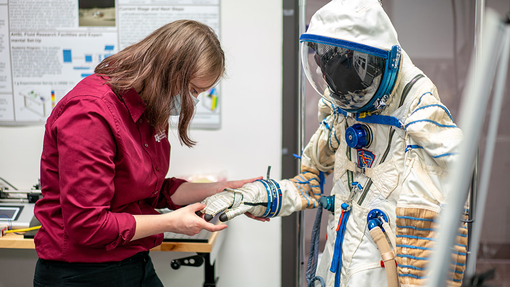 女学生站在一件宇航服前，拿着宇航服上的手套向下看。