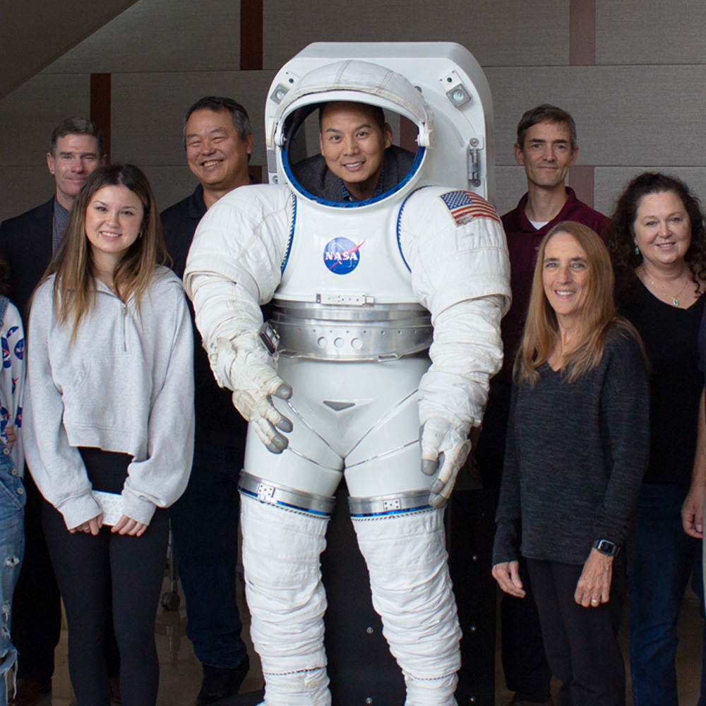 A group of NASA representatives, their families and Texas A&M faculty stand together around a life-sized space suit model and smile at the camera.