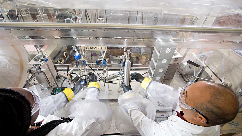 男女研究人员穿着实验室大褂和其他安全装备，在一个封闭的实验站工作