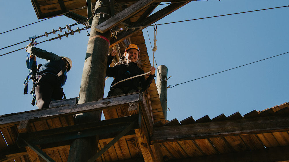 一位年轻女子站在塔顶，在滑索前对着镜头微笑。