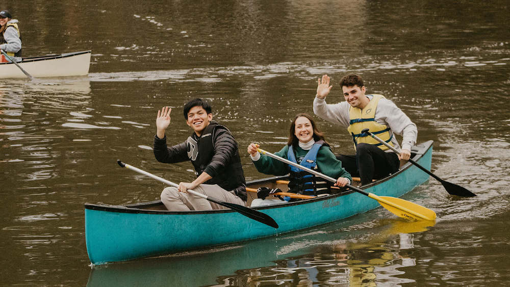 三个学生在湖上的独木舟上对着镜头挥手。