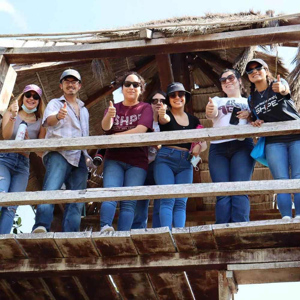 Seven students doing a gig'em from inside wooden hut with straw roof