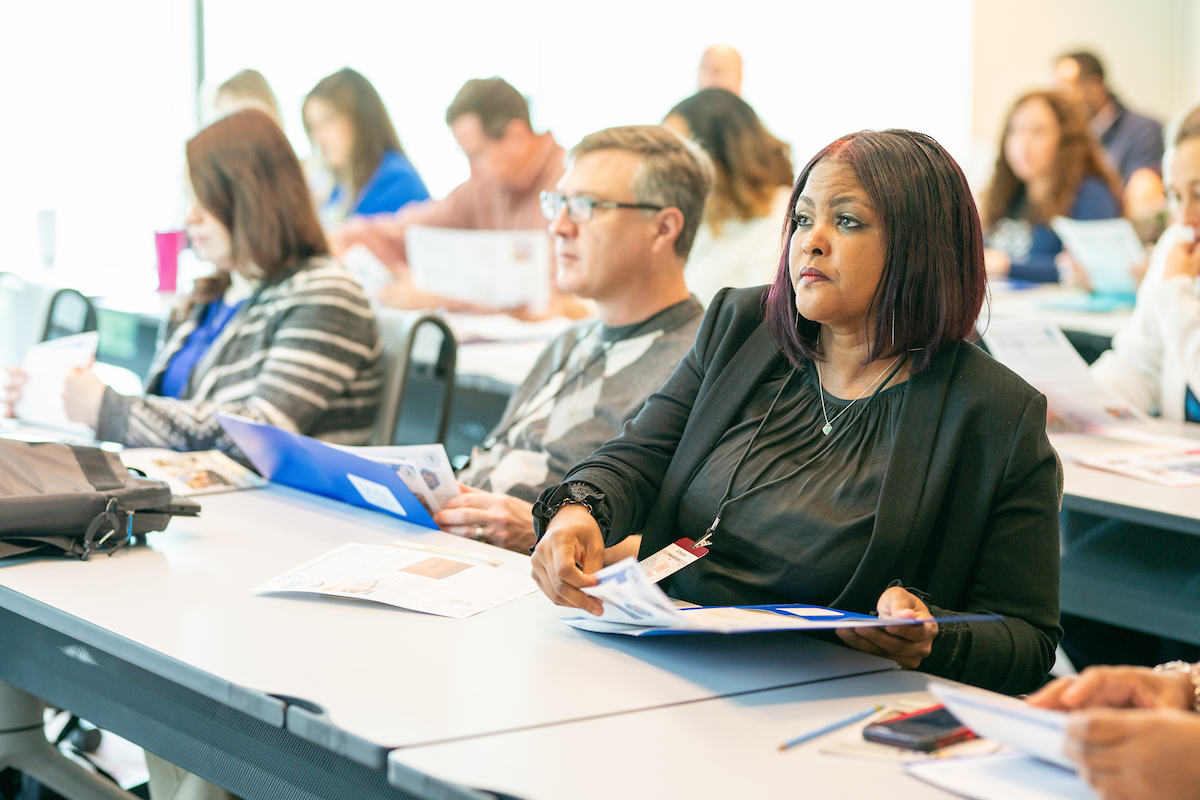 A woman sits in a full classroom in business attire pulling papers out of a folder while listening to a speaker.