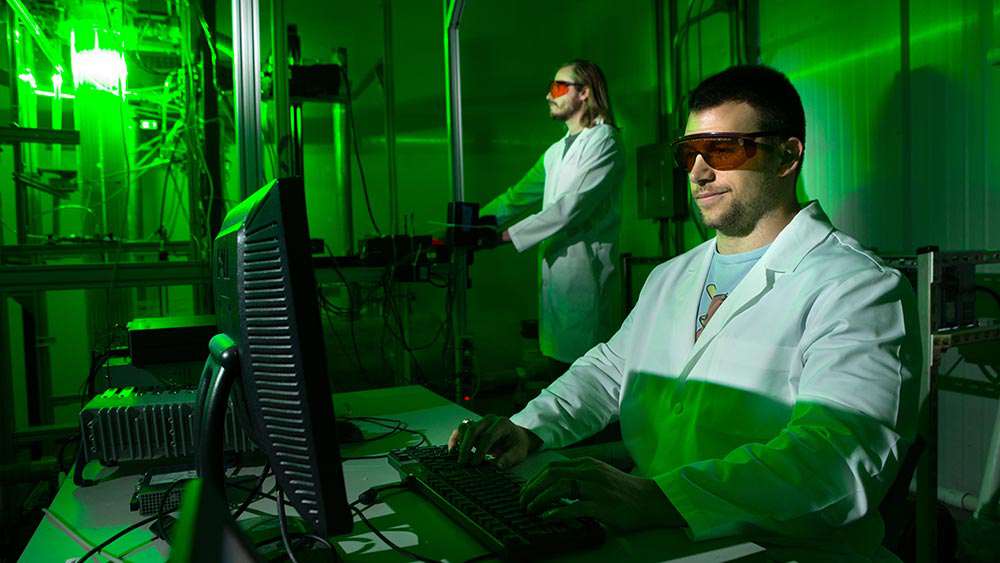 Two grad students in scientific gear are lit by the green glow of a reactor.